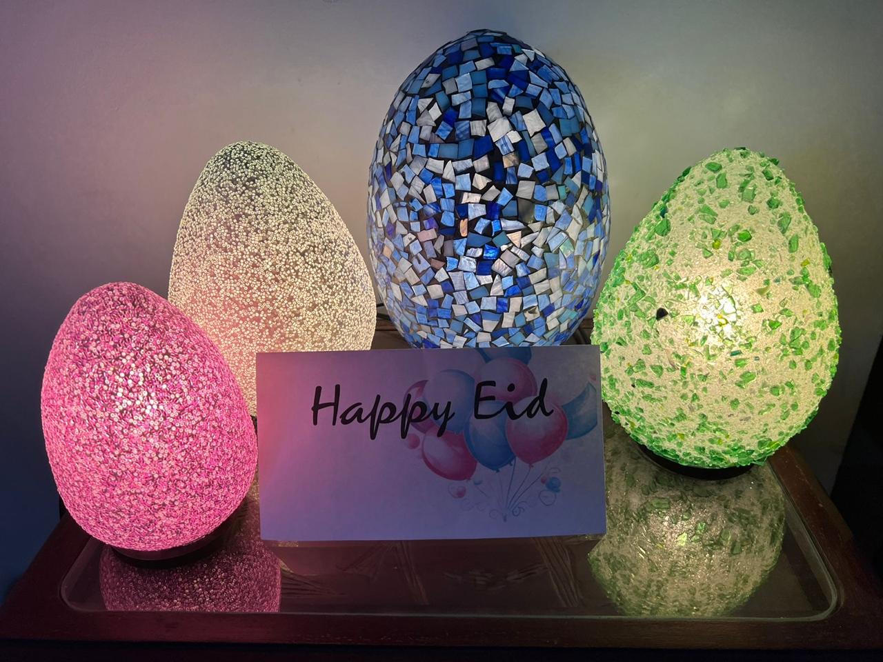 Decorative mosaic eggs with 'Happy Eid' card on a reflective surface